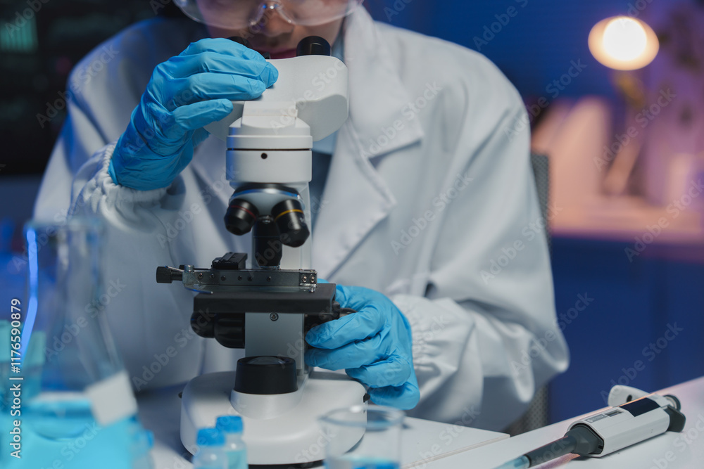 Scientist wearing protective gear adjusts a microscope, conducting research in a modern laboratory filled with scientific equipment and bathed in blue light Scientist wearing protective gear adjusts a microscope, conducting research in a modern laboratory filled with scientific equipment and bathed in blue light
