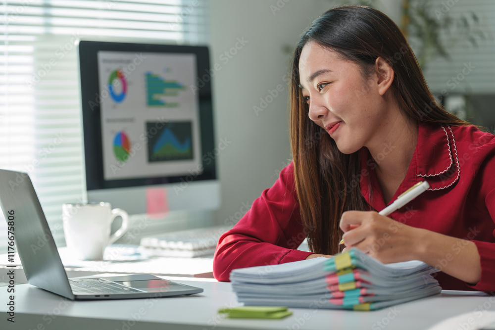 Young asian businesswoman in red shirt taking notes and working on a laptop in a modern office with charts displayed on a desktop computer screen Young asian businesswoman in red shirt taking notes and working on a laptop in a modern office with charts displayed on a desktop computer screen