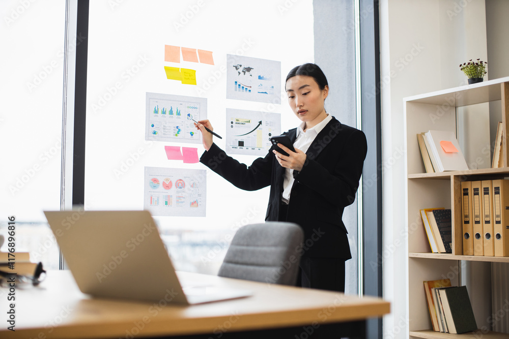 Naklejka premium Asian businesswoman in black suit presenting data on transparent board in modern office. She uses smartphone while engaging with charts, reflecting professional and focused work environment.