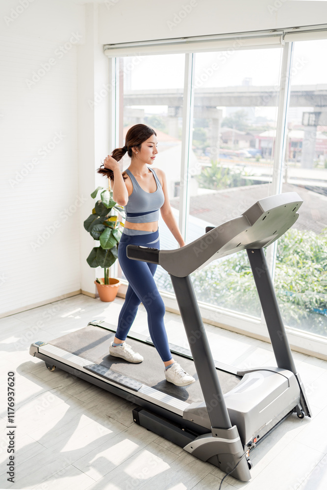 Fototapeta premium A young Asian woman is walking on a treadmill in a bright, modern room with large windows and a potted plant nearby. Indoor working out or exercise at home, wearing sportswear.