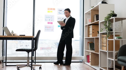 Wallpaper Mural Asian businesswoman stands in office reviewing reports on tablet. Professional attire. Office space with bookshelves and charts. Corporate environment suggests productivity and focus. Torontodigital.ca