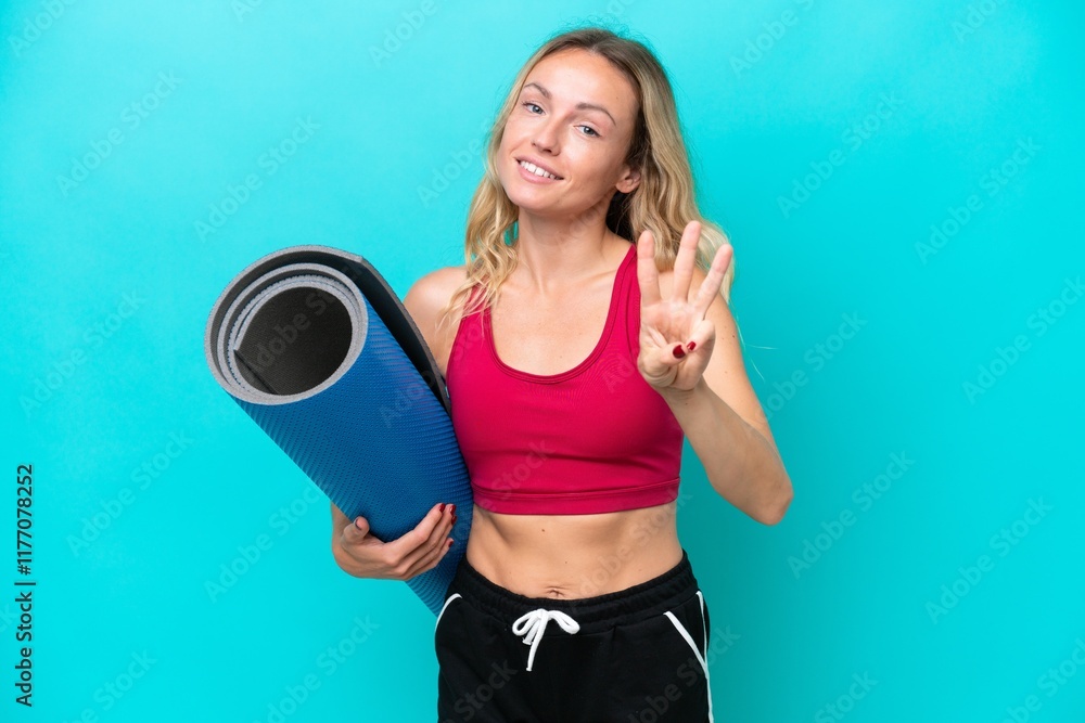Young sport Russian woman going to yoga classes while holding a mat isolated on blue background happy and counting three with fingers Young sport Russian woman going to yoga classes while holding a mat isolated on blue background happy and counting three with fingers