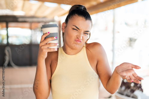 Free Download Young pretty woman holding a take away coffee at outdoors making doubts gesture while lifting the shoulders - GFXLoad
