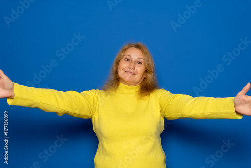Caucasian woman in her 40s wearing a yellow sweater stretching out her hands, hugging, extending her arms to hug, embracing someone, standing against a blue background