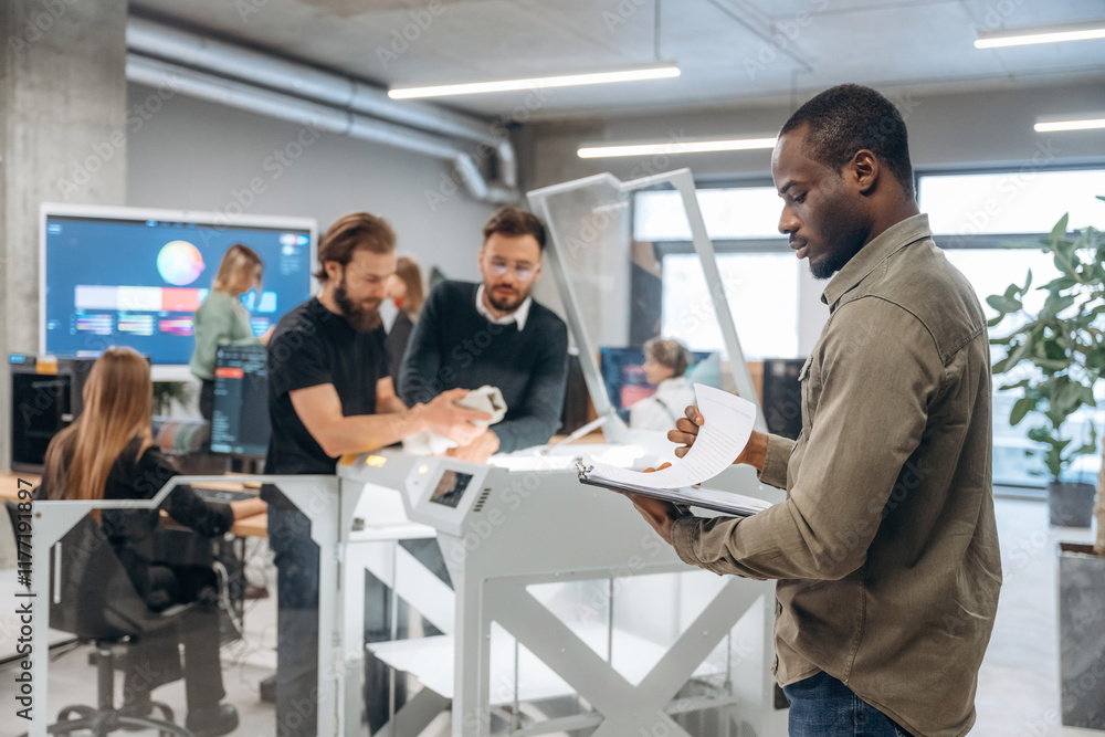 African American man is standing in front of his colleagues. People are in the office by the 3d printer African American man is standing in front of his colleagues. People are in the office by the 3d printer