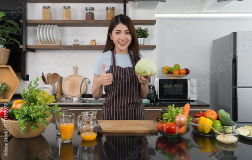 Young asian housewife dressed in an apron raise finger thumb up while holding fresh lettuce. Morning atmosphere in a modern kitchen. photo