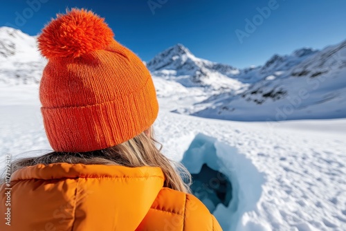 A hiker in an orange jacket stands before an ice cavity, gazing at magnificent snowy mountains, symbolizing exploration and the thrill of nature's wonders. photo