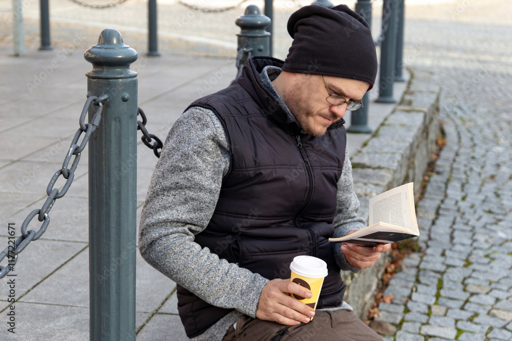 A man reading a book outdoors in an autumn city.