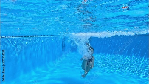 A girl jumps doing a cannonball into the swimming pool and dive under the water with a bright, clear blue background