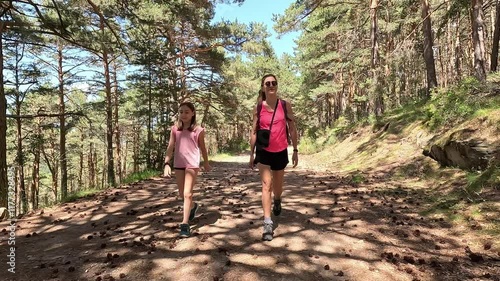 Mother and her daughter hikers talking while walking on a forest trail through tall pine trees on a sunny summer day, with dappled sunlight filtering through the canopy, in the Canencia mountain, the 