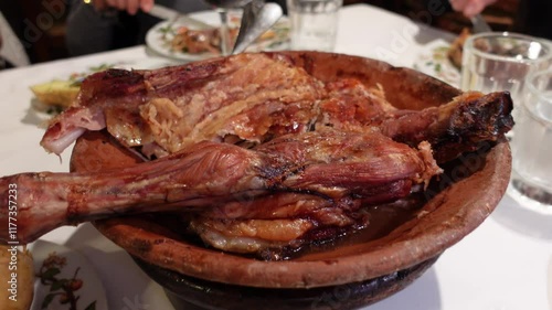Roasted lamb served in a traditional clay dish with baked potatoes and bread on a dining table, accompanied by red wine and a cozy atmosphere