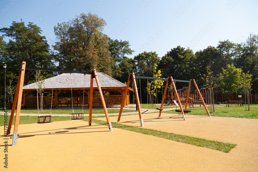 Fototapeta premium Children's playground with swings and a wooden gazebo. Sunny day, lush green trees in background.