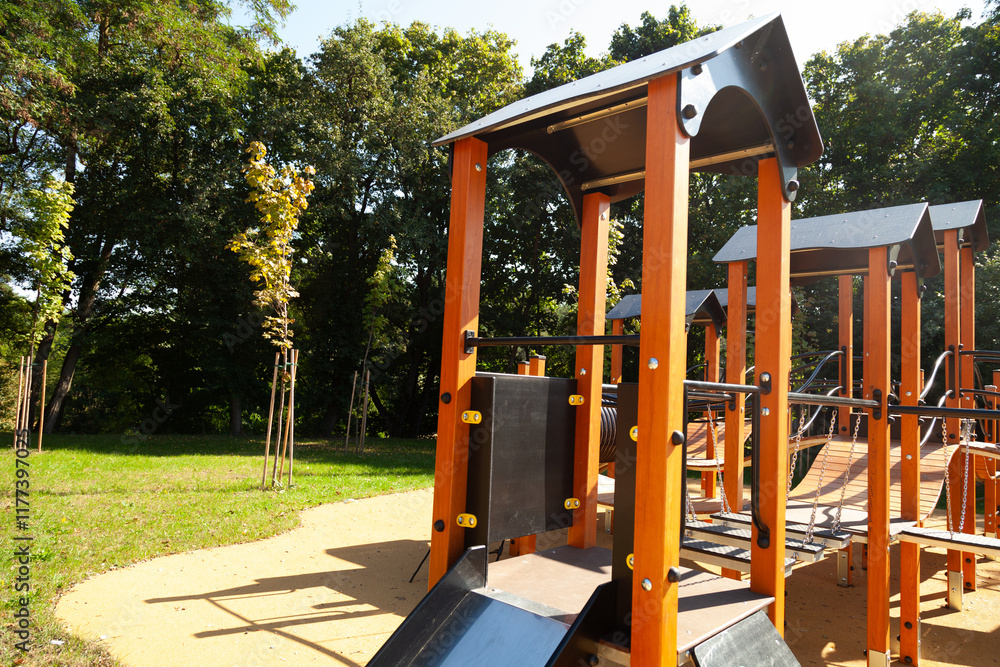 Modern playground equipment in a park setting on a sunny day.  Children's play structure with orange wood and black accents.