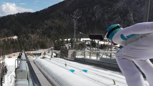 Rear view of an athlete practicing ski jumps, view from the board, an inrun border, on a track and mountains, static shot.
