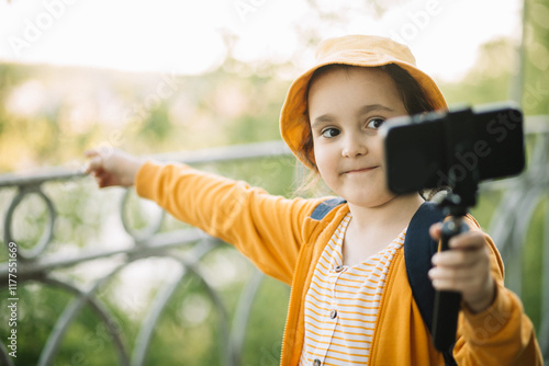 Cute little girl taking selfie with mobile phone looking at camera in a park. Kid blogger recording video vlog. photo