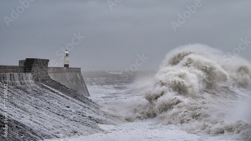 A huge winter storm hitting the coast of the UK. Giant waves crash into a breakwater, with a lighthouse. The sky is grey and moody photo