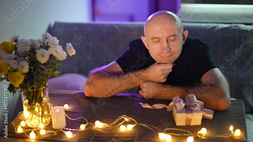 Man seated at table contemplating wrapped gift, flowers nearby, soft fairy lights illuminating intimate scene