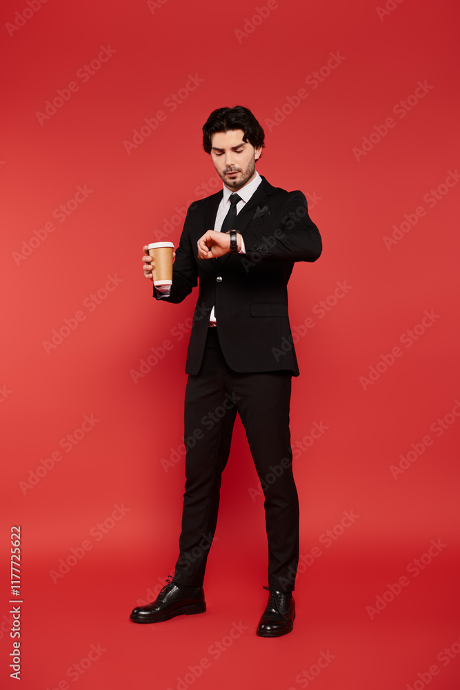 Young man in stylish suit checks watch while holding coffee against bright red backdrop