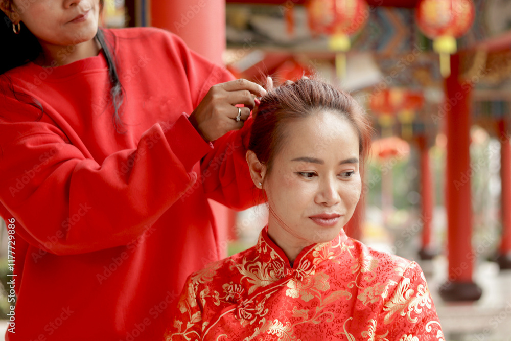Asian women with red traditional cheongsam qipao dress styled her hair by her friend while sit in Chinese building. Happy people celebrating Chinese New Year. Chinese tradition festival holiday. Asian women with red traditional cheongsam qipao dress styled her hair by her friend while sit in Chinese building. Happy people celebrating Chinese New Year. Chinese tradition festival holiday.