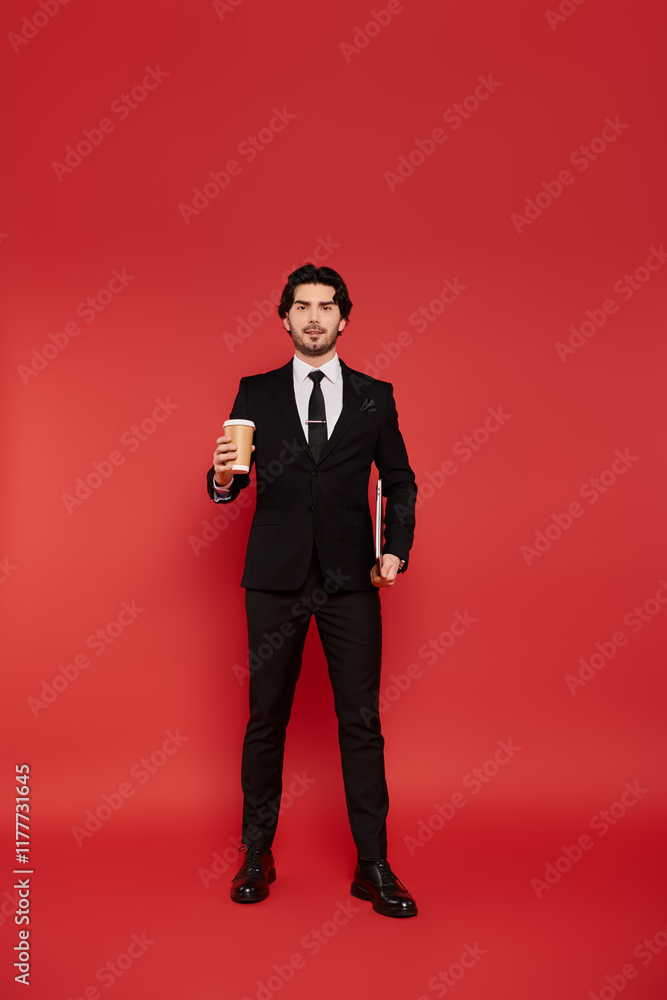 Young man in black suit holding coffee while standing confidently against bright red backdrop Young man in black suit holding coffee while standing confidently against bright red backdrop