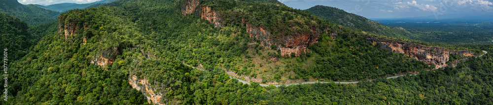Aerial panoramic view of lush green hills and valleys intersected by a winding mountain road. The landscape features rugged cliffs, dense forests, and a distant horizon under a partly cloudy sky. Aerial panoramic view of lush green hills and valleys intersected by a winding mountain road. The landscape features rugged cliffs, dense forests, and a distant horizon under a partly cloudy sky.