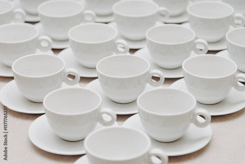 white empty porcelain cups on saucers with spoons in several rows on white tablecloth, serving tables for banquet in restaurant or cafe, pattern, background white empty porcelain cups on saucers with spoons in several rows on white tablecloth, serving tables for banquet in restaurant or cafe, pattern, background