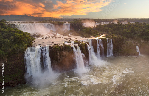 Obraz na płótnie Majestic Iguazu Falls with powerful cascades flowing amidst lush green vegetation, illuminated by a vibrant sunset sky w salonie