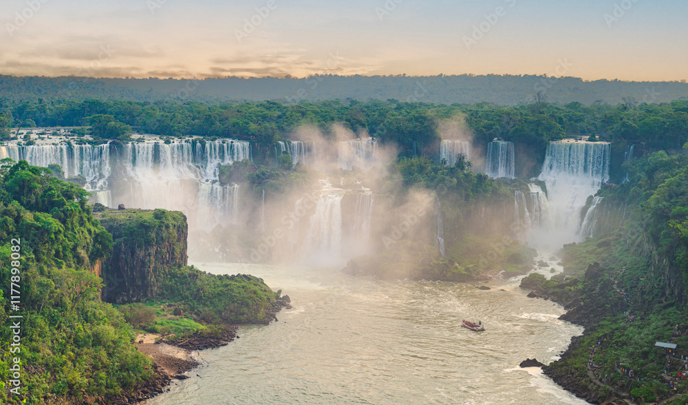 View of the Iguazu Falls surrounded by lush green forest under a vibrant orange sky at sunset, with boats navigating the river below