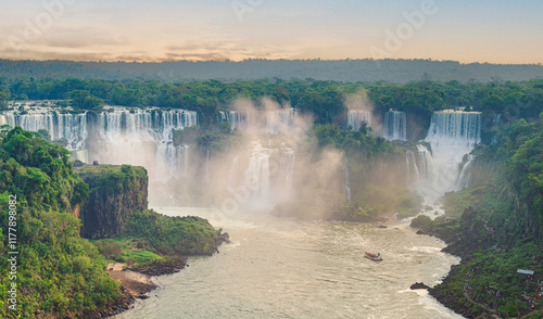 Obraz na płótnie View of the Iguazu Falls surrounded by lush green forest under a vibrant orange sky at sunset, with boats navigating the river below w salonie