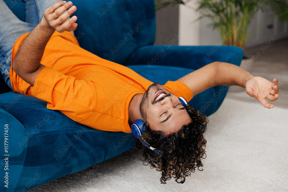 © ViDi Studio - Side view young Indian man wear orange casual clothes lay down on blue sofa couch listen music in headphones stay home hotel flat rest relax spend free spare time in living room indoor Lounge concept