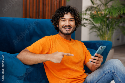 Young Indian man wear orange casual clothes sit near blue sofa couch hold use point on mobile cell phone stay at home hotel flat rest relax spend free spare time in living room indoor. Lounge concept. photo