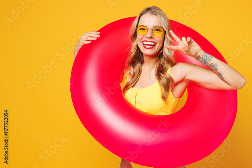 Young beautiful woman wears bikini swimsuit sunglasses rest near hotel pool hold pink rubber ring show cover eye with v-sign isolated on plain yellow background. Summer vacation sea sun tan concept.