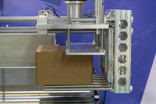 Photos Close-up view of modern heavy robotic arm manipulator moving large blank cardboard box on conveyer belt in industrial building