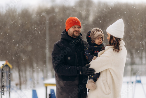 A young couple enjoys the winter chill as they walk through a snowy street with their child in a stroller. Snowflakes fall around them, creating a picture-perfect moment of joy photo