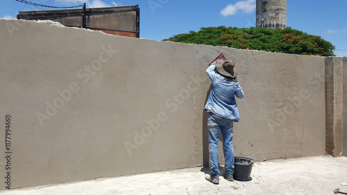 Pedreiro de roupa azul e chapéu preto, fazendo acabamento de construção do muro de cimento. photo