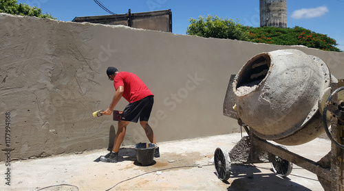 Pedreiro realizando trabalho de acabamento na construção do muro de cimento, e betonera ao lado com massa de cimento. photo