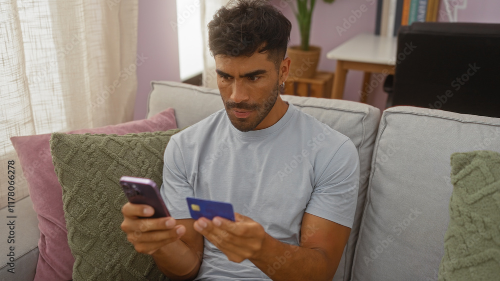 Young man sitting at home using smartphone and credit card appearing focused and thoughtful in the living room interior setting Young man sitting at home using smartphone and credit card appearing focused and thoughtful in the living room interior setting