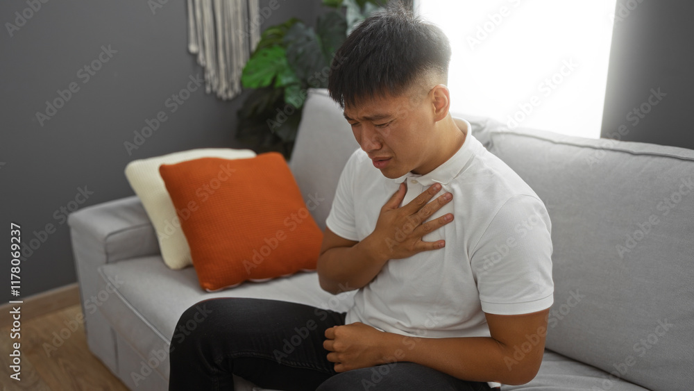 Young man experiencing chest discomfort while seated in a living room with cushions and a gray sofa. Young man experiencing chest discomfort while seated in a living room with cushions and a gray sofa.