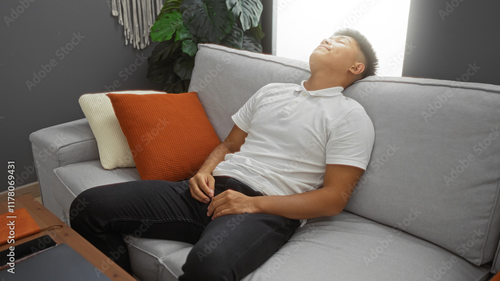 Young man relaxing on a couch in a living room at home with pillows and plants in the background Young man relaxing on a couch in a living room at home with pillows and plants in the background