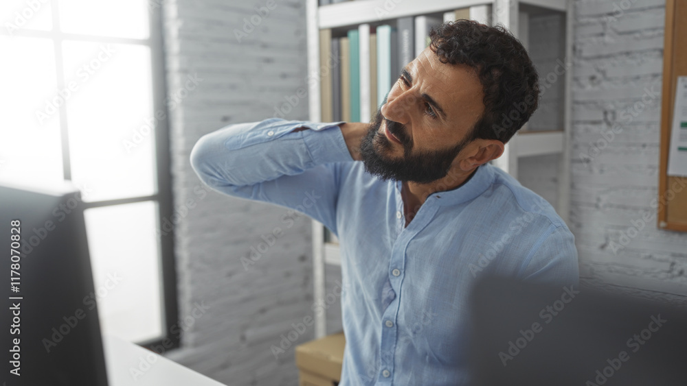 Fototapeta premium Young man with beard and blue shirt stretching his neck in a bright office, surrounded by books and documents, suggesting a professional and focused work environment.