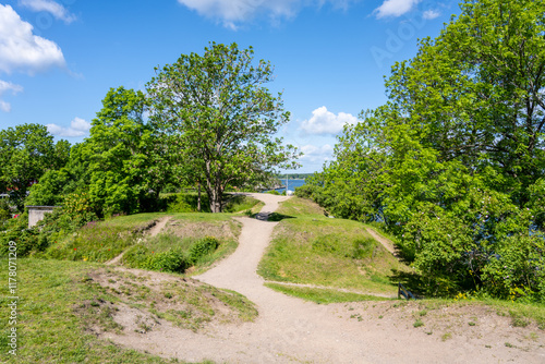 A tranquil pathway winds through lush greenery at Battery Park on Vaxholm Island near Stockholm. Visitors enjoy outdoor activities surrounded by vibrant nature and scenic views of the water. photo