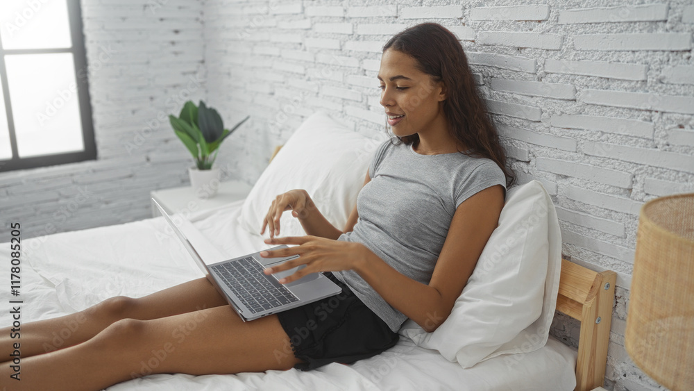 Young woman relaxing on bed in modern bedroom using laptop with white brick walls and green plant next to window Young woman relaxing on bed in modern bedroom using laptop with white brick walls and green plant next to window
