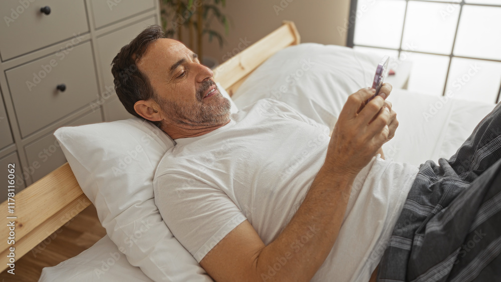 Obraz premium Middle-aged hispanic man lying on a bed in a modern bedroom, engaged with his smartphone, providing a casual glimpse into a relaxed home environment.