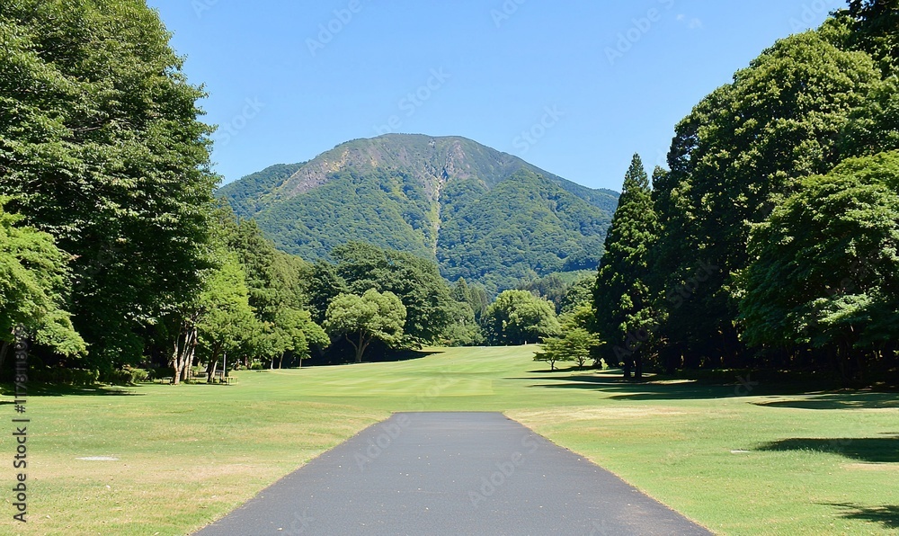 Fototapeta premium Paved path through green golf course towards forested mountain under blue sky.
