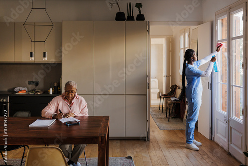 Caregiver cleaning windows while senior woman sitting on dining table photo