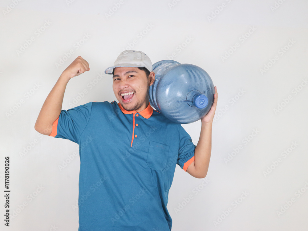 A happy Asian delivery courier is carrying a gallon or big water bottle of mineral water with a pose showing bicep muscles. raising his fist. A happy Asian delivery courier is carrying a gallon or big water bottle of mineral water with a pose showing bicep muscles. raising his fist.