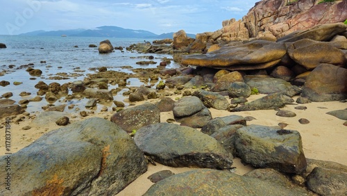 A rocky coastal landscape with scattered boulders in Hon Chong cape rocks garden on the Nha Trang beach, Vietnam with a calm sea and mountains in background.