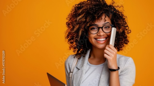 A young woman with curly hair smiling while talking on her phone and holding a laptop, posed against a vibrant orange background.

