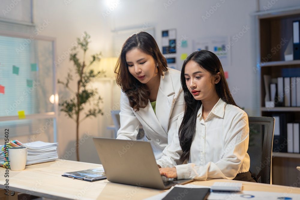 Two young asian businesswomen working late in a brightly lit office, collaborating on a project while using a laptop and engaging in a focused discussion about their goals Two young asian businesswomen working late in a brightly lit office, collaborating on a project while using a laptop and engaging in a focused discussion about their goals