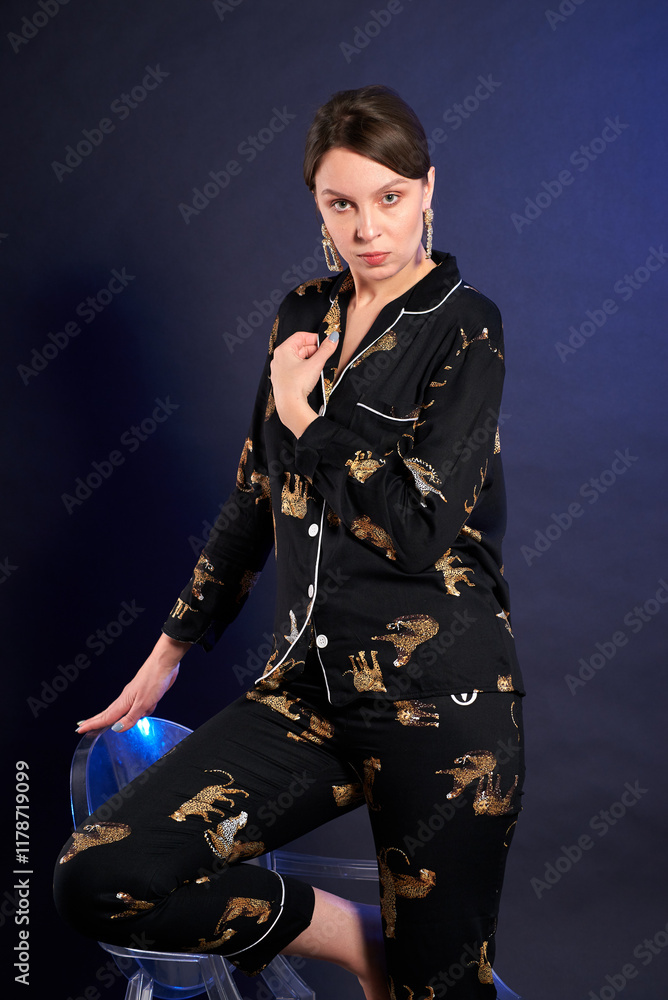 Naklejka premium A woman in a black and gold pajama set poses for the camera in a studio against a dark background. The outfit features animal prints, giving it a playful and whimsical look. Confident pose of a woman